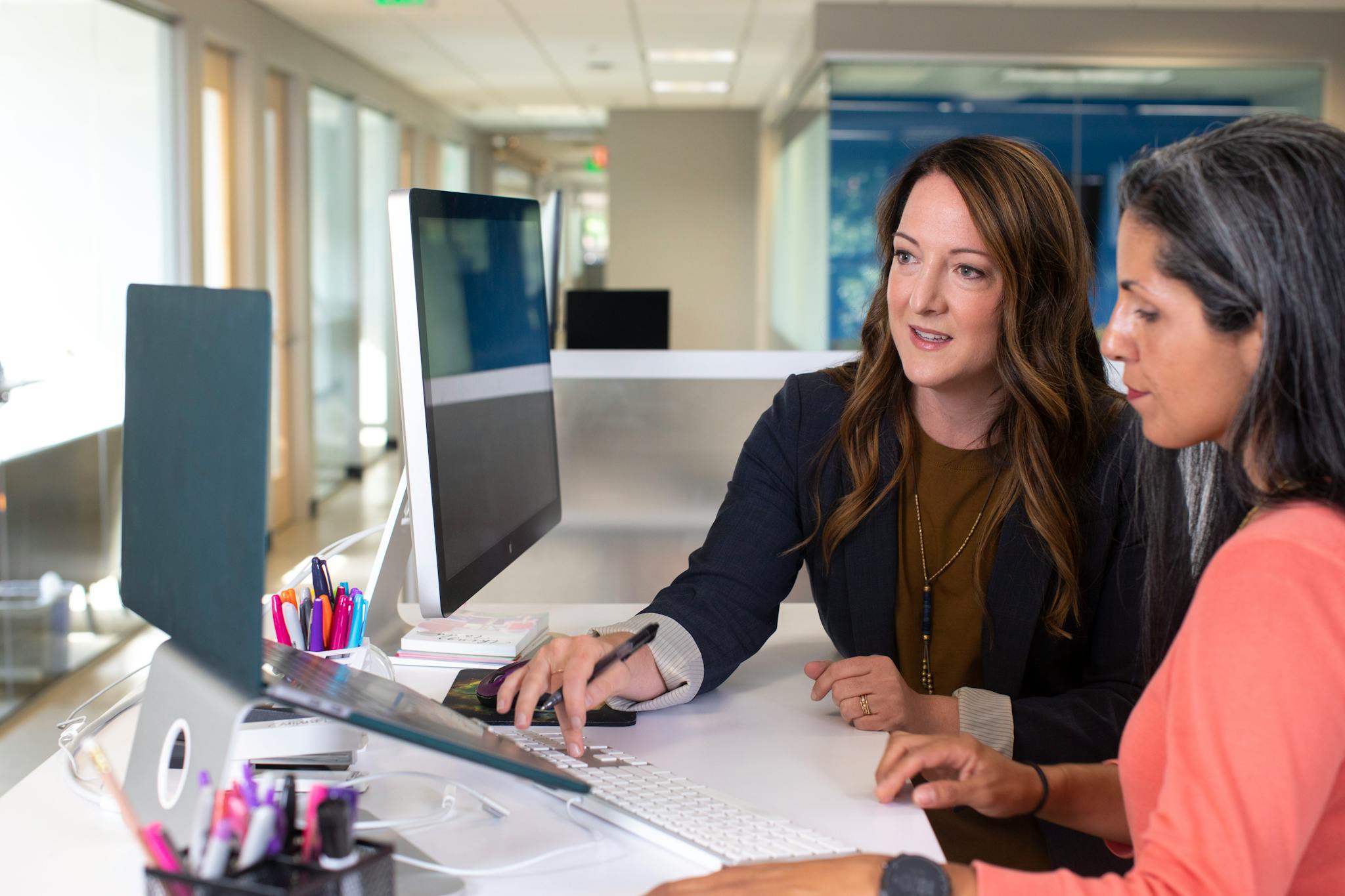 Two women working together at a desk with computers in a bright office space.